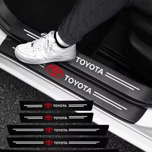 May include: Black carbon fibre car door sill protectors featuring the Toyota logo in red and the word "TOYOTA" in white. The protectors have white accent lines and are shown installed in a car.
