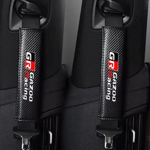 May include: Two black seat belt covers with a carbon fiber pattern. Each cover has the text "GR Gazoo Racing" in white and red. The covers are attached to black seat belts in a car interior.