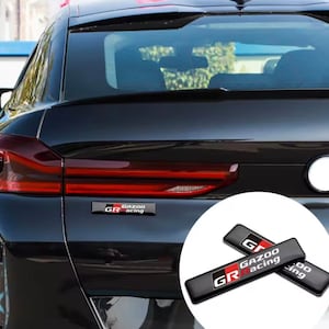 May include: Black car with red taillights and two black and red GR Gazoo Racing emblems. The emblems are rectangular with the letters "GR" and the words "Gazoo Racing" in white.