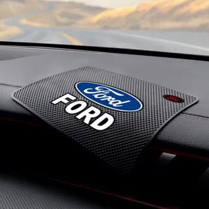 May include: Black textured car dashboard mat featuring the Ford logo in white and blue. The word "FORD" is also displayed in large white letters. A small red accent is visible on the mat.