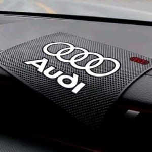 Può includere: Un tappetino antiscivolo nero e rettangolare per cruscotto, caratterizzato da una superficie testurizzata con il logo e la scritta Audi stampati in bianco al centro. Il tappetino è posizionato su un cruscotto scuro, con un piccolo accento rosso.