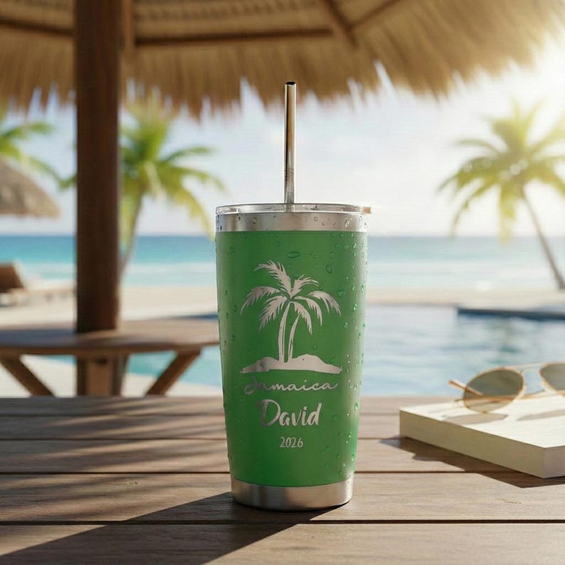 May include: A green travel tumbler with a silver lid and straw. The tumbler features a palm tree design with the word "Jamaica" and the name "David" and the year "2026". The tumbler is sitting on a wooden surface near a pool and beach.