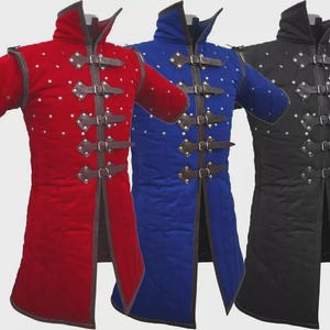 Può includere: Tre tuniche medievali imbottite, rosse, blu e nere. Ogni tunica presenta un colletto alto, maniche corte e cinturini in pelle con fibbie. Borchie argentate sono uniformemente distanziate sul davanti.
