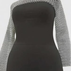 Puede incluir: Una camisa de cota de malla plateada con mangas largas exhibida en un maniquí negro. La cota de malla está hecha de pequeños anillos de metal entrelazados. El maniquí es negro y tiene un diseño simple y elegante. El fondo es blanco.