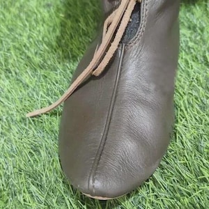 Puede incluir: Primer plano de un zapato de cuero marrón con diseño de cordones. El zapato es de color marrón oscuro y parece estar hecho de cuero. Los cordones son de un marrón más claro. El zapato está sobre una superficie verde.