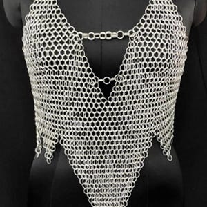 Może przedstawiać: Srebrny top z kolczugi na czarnym manekinie. Top w stylu halter ma dekolt w kształcie litery V i trójkątny dół. Na szyi manekina widnieje numer "12". Kolczuga wykonana jest z małych, zazębiających się metalowych pierścieni.