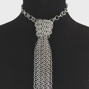 Peut inclure: Un collier en cotte de mailles argenté avec un design en forme de cravate. Le collier présente une chaîne ras du cou qui se connecte à une section triangulaire, qui se transforme en une longue frange rectangulaire en cotte de mailles. La pièce est présentée sur un mannequin noir.