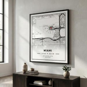 Może przedstawiać: Oprawiona czarno-biała mapa Miami z czerwonym sercem oznaczającym lokalizację. Mapa zawiera imiona "JANE ♥ ADAM" oraz datę "06.05.2022". Nazwa miasta "MIAMI" jest wyśrodkowana na dole.