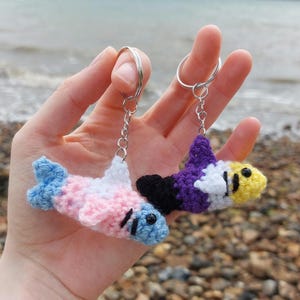 May include: Two crocheted fish keyrings. One is pink, white, and blue, the other is purple, black, white, and yellow. Each keyring has a silver ring and chain. The background is a blurry view of a beach.