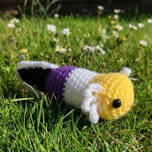 May include: A crocheted plush toy shaped like an axolotl, featuring a yellow head with white external gills, a black eye, and a body striped in white, purple, and black. The soft, handmade creature has a rounded head and a long, segmented tail.
