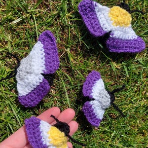 May include: Handmade crocheted butterfly decorations in purple, white, grey, and yellow, with black antennae. The butterflies are arranged on green grass. The butterflies are a craft item.