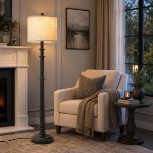 May include: A tall floor lamp with a dark charcoal gray metal post and a cylindrical beige fabric drum shade. The lamp features a round base and a slender, tiered vertical stand that supports the illuminated shade, providing warm ambient light.
