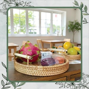 May include: A round, woven wicker tray holds a vibrant pink dragon fruit, packaged macarons, and a small box. Another tray contains lemons and oranges. The trays sit on a wooden table in a bright room.