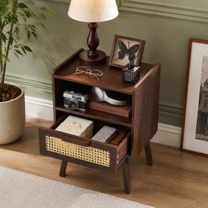 May include: A dark brown wooden nightstand featuring an open shelf and a single pull-out drawer with a light-colored woven rattan front panel. The nightstand is supported by four tapered legs and contains various personal items on its surfaces.