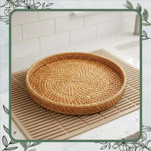 May include: A round, woven rattan tray in natural tones sits on a beige drying mat. The tray's intricate weave creates a textured surface, perfect for serving or displaying items. The neutral colors complement various kitchen styles.