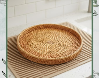 May include: A round, woven rattan tray in natural tones sits on a beige drying mat. The tray's intricate weave creates a textured surface, perfect for serving or displaying items. The neutral colors complement various kitchen styles.