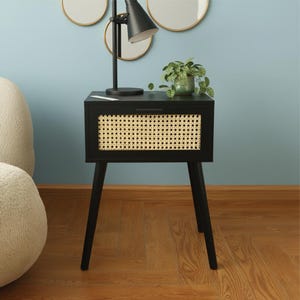 May include: A black nightstand with a woven rattan front panel and tapered legs. A small black lamp and a potted plant sit on top of the nightstand. The nightstand is placed on a wooden floor.