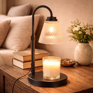 May include: A black candle warmer lamp with a clear, textured glass shade. A lit candle sits on the base of the lamp. The lamp is on a wooden surface with a vase of flowers and books in the background.