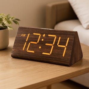 May include: A triangular digital alarm clock with a dark brown wood grain finish. The front face displays the time 12:34 in bright orange LED numerals, with an AM indicator and a small bell icon visible above the numbers on the clock display.