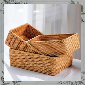 May include: Three rectangular woven rattan baskets in varying sizes are stacked. The baskets are a warm, natural tan color, with a tightly woven pattern. The image is well-lit, highlighting the texture and craftsmanship of the storage containers.