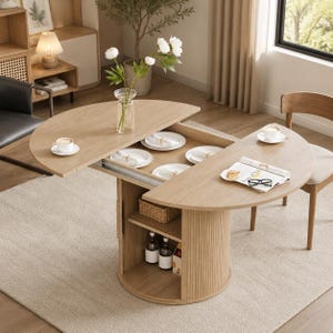 May include: A light oak extendable dining table with a semi-circular top and a central cylindrical base featuring vertical slats. The table includes integrated storage shelves, white ceramic plates, a glass vase with white flowers, and a magazine.