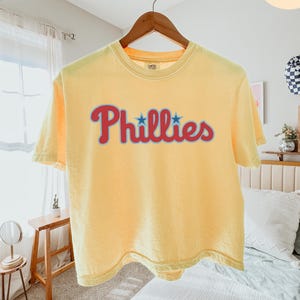 May include: A butter yellow short-sleeve t-shirt featuring the word Phillies in red script with a light blue outline and two small blue stars above the letters. The shirt is displayed on a wooden hanger against a neutral indoor setting.