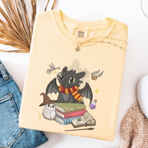 May include: A pale yellow t-shirt featuring a graphic of a black dragon wearing round glasses and a red and yellow striped scarf. The dragon sits atop a stack of books next to a white owl, a brown wizard hat, a wand, a potion, and a flying key.