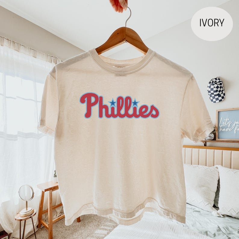 May include: An ivory short-sleeve t-shirt featuring the word Phillies printed in a red script font with blue outlines and two blue stars above the letters. The shirt is displayed on a wooden hanger against a light-colored interior background.