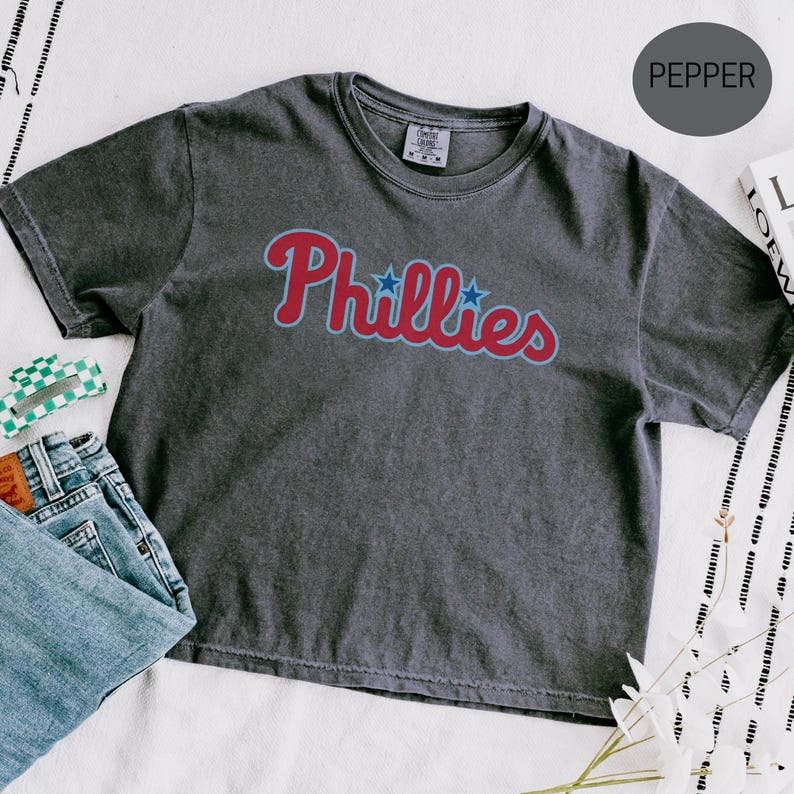 May include: A charcoal grey short-sleeve t-shirt with a vintage-wash finish. The front features the word Phillies in a red script font with a light blue outline and two small blue stars replacing the dots above the letter i.