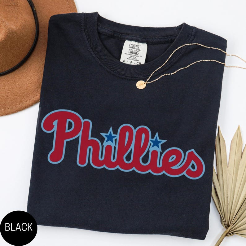 May include: A dark navy blue short-sleeve t-shirt featuring the word Phillies in a red script font with a light blue outline and two blue stars replacing the dots on the letter i. The shirt has a Comfort Colors brand label at the inner neckline.