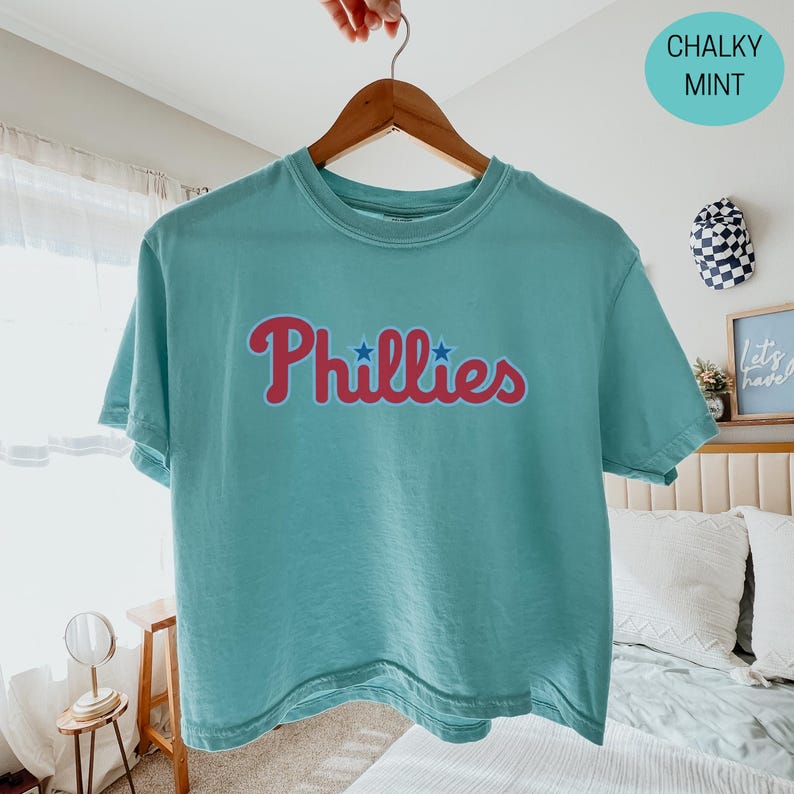 May include: A chalky mint green short-sleeve t-shirt featuring the word Phillies in a red script font across the chest. The text includes two small blue stars positioned above the letters i. The shirt is displayed on a wooden hanger.