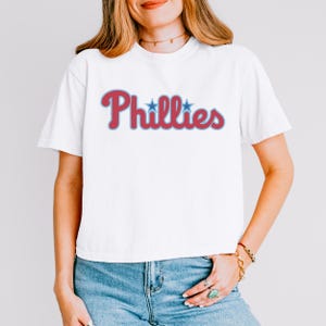May include: A white short-sleeve t-shirt featuring the word Phillies printed in a red script font with a light blue outline. Two small light blue stars are positioned above the letters i in the word. The shirt has a crew neckline.