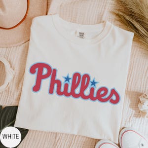 May include: A white short-sleeved t-shirt featuring the word Phillies printed in a red script font with a light blue outline. Two small blue stars are positioned above the letters i and l. The shirt has a crew neckline and a Comfort Colors label.