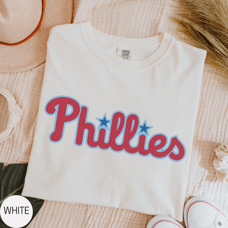 May include: A white short-sleeved t-shirt featuring the word Phillies printed in a red script font with a light blue outline. Two small blue stars are positioned above the letters i and l. The shirt has a crew neckline and a Comfort Colors label.