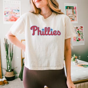 May include: A cream-colored short-sleeve t-shirt featuring the word Phillies printed in a red script font with a light blue outline. Two small blue stars are positioned above the letters i in the word. The shirt has a crew neckline and a relaxed fit.