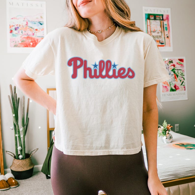 May include: A cream-colored short-sleeve t-shirt featuring the word Phillies printed in a red script font with a light blue outline. Two small blue stars are positioned above the letters i in the word. The shirt has a crew neckline and a relaxed fit.