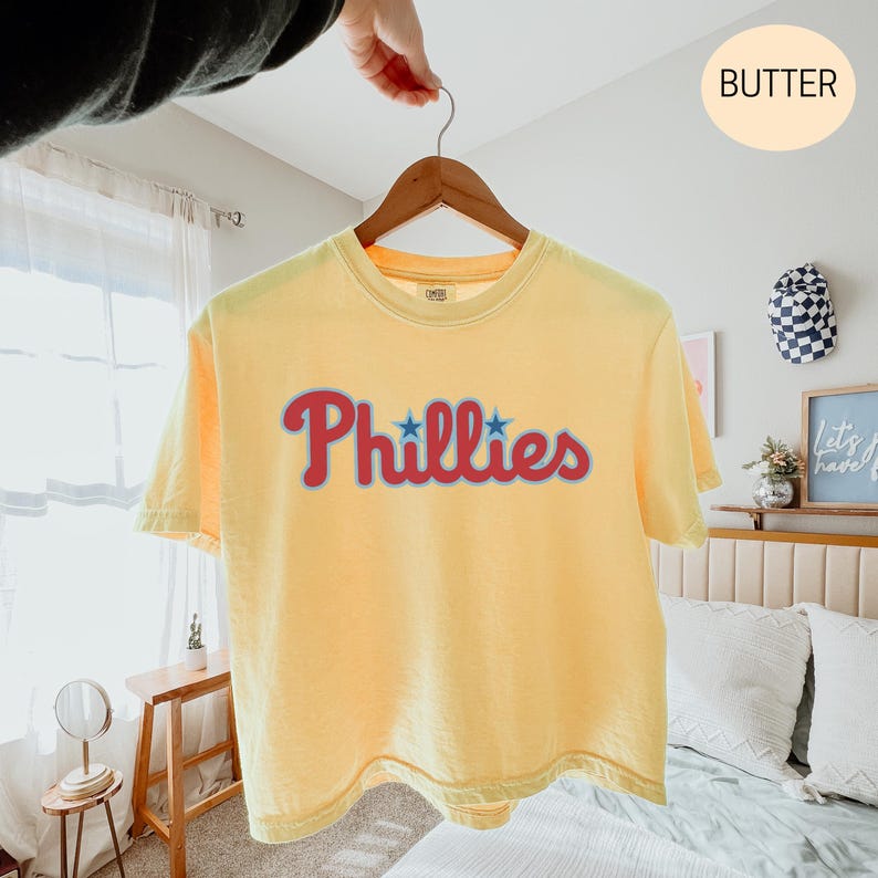 May include: A butter yellow short-sleeve t-shirt featuring the word Phillies in red script with a light blue outline and two small blue stars above the letters. The shirt is displayed on a wooden hanger against a neutral indoor setting.