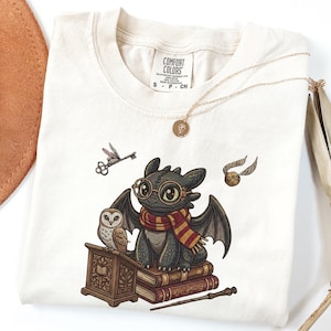 May include: An off-white t-shirt featuring a graphic of a black dragon wearing round glasses and a red and yellow striped scarf, perched on a stack of books next to an owl, a wooden lectern, a flying key, a golden snitch, and a wand.