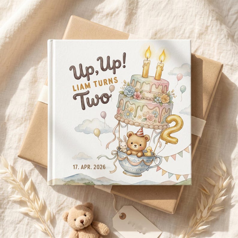 Puede incluir: Un libro de tapa dura cuadrado con cubierta blanca que presenta una ilustraci&oacute;n en acuarela de un osito de peluche, un conejo y un patito en un globo aerost&aacute;tico de taza de t&eacute;. Un pastel de pisos con dos velas est&aacute; encima. Texto: Up, Up! LIAM TURNS Two 17. APR. 2026.