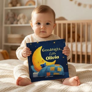 May include: A baby holds a children's book titled "Goodnight, Little Olivia." The book cover features a smiling crescent moon reading a book under a starry sky. The baby is wearing a cream-colored sweater and sitting on a bed.