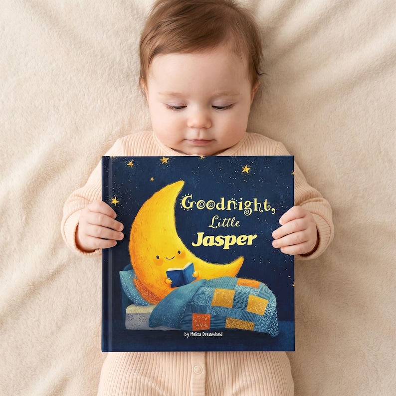 May include: A baby holds a children's book titled "Goodnight, Little Jasper." The book cover features a smiling yellow moon reading in bed under a starry night sky. The book is by Melisa Dreamland.