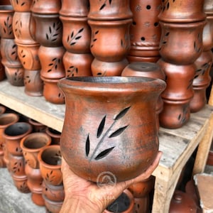 May include: A terracotta-colored ceramic planter with a leaf-shaped cutout design. The planter is round with a wide opening and is held in a person's hand. Other similar planters are stacked in the background.