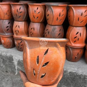 May include: Terracotta-colored ceramic flower pots with leaf-shaped cutouts. The pots are stacked, showcasing a rustic design. The pots have a slightly flared rim and a textured surface. The image includes the text "ARTA TERRA".