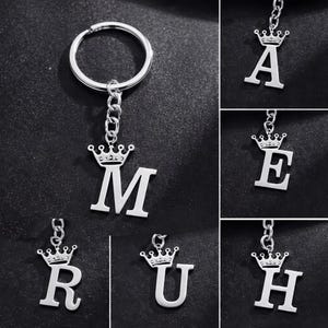 Puede incluir: Llaveros de metal plateado con letras del alfabeto, cada uno coronado con un diseño de corona. Los llaveros se muestran sobre un fondo oscuro. Las letras incluyen M, A, E, R, U y H.