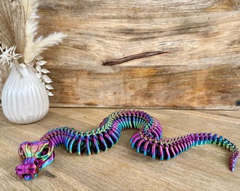 May include: A colorful, articulated snake toy in shades of purple, blue, green, and gold. The snake is posed on a wooden surface, with a white vase and dried flowers in the background. The snake's design is detailed and eye-catching.
