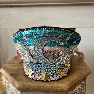 May include: A blue and silver sequined hat with a crescent moon design and a pink velvet band. The hat is decorated with beads and rhinestones.