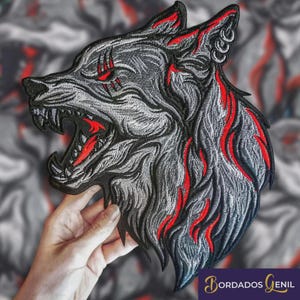 Puede incluir: Parche bordado de cabeza de lobo en tonos grises y negros con detalles rojos. El lobo está gruñendo, con la boca abierta, y tiene detalles rojos alrededor de los ojos y en el pelaje. Para ropa o accesorios.