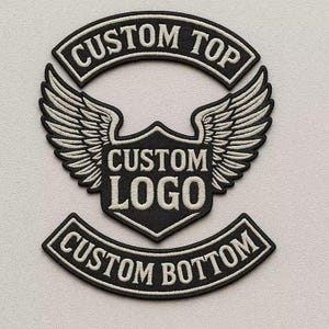 以下が含まれることがあります： 「CUSTOM TOP」、「CUSTOM LOGO」、および「CUSTOM BOTTOM」のテキストが刺繍された白と黒のパッチ。中央のパッチは、様式化された翼を持つシールド形状を特徴としています。