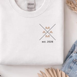 Peut inclure: Sweat-shirt blanc avec le mot "MAMA" dans un design en croix, avec le texte "est. 2026" en dessous. Les lettres sont de couleur beige. Le sweat-shirt est plié.