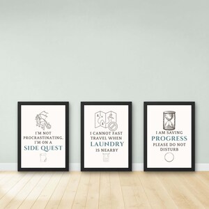 Op de afbeelding: Drie ingelijste prints met zwarte randen en witte achtergronden. Elke print heeft een ander ontwerp en tekst: een draak, een kaart en een zandloper. De tekst bevat zinnen als "Side Quest", "Laundry" en "Progress".